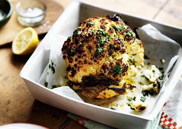 Whole head of roasted cauliflower, with a scorched top, scattered with parsley, in a square roasting tray.