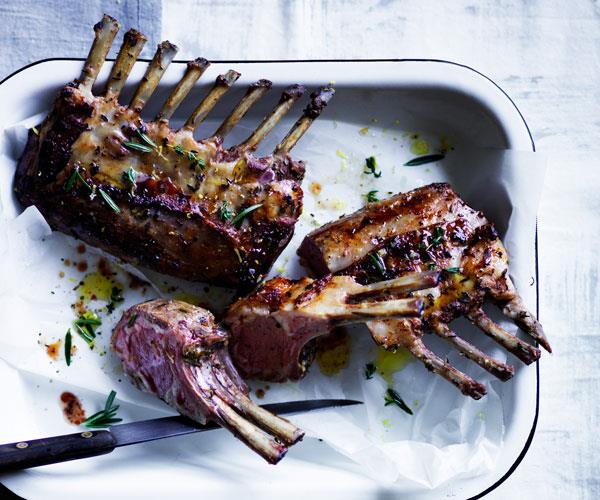 A roast rack of lamb, scattered with rosemary, on a white roasting tray.