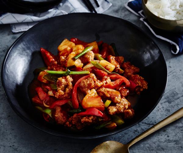 A black bowl holding sweet and sour pork with sliced capsicum and chunks of pineapple