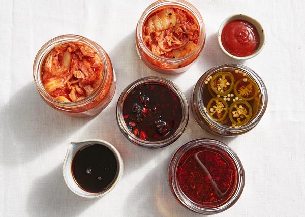 Over the top shot of jars containing kimchi, teriyaki sauce, textural XO sauce, Korean hot sauce and pickled pickled jalapeños o a white surface. The jars cast long shadows on the surface. 