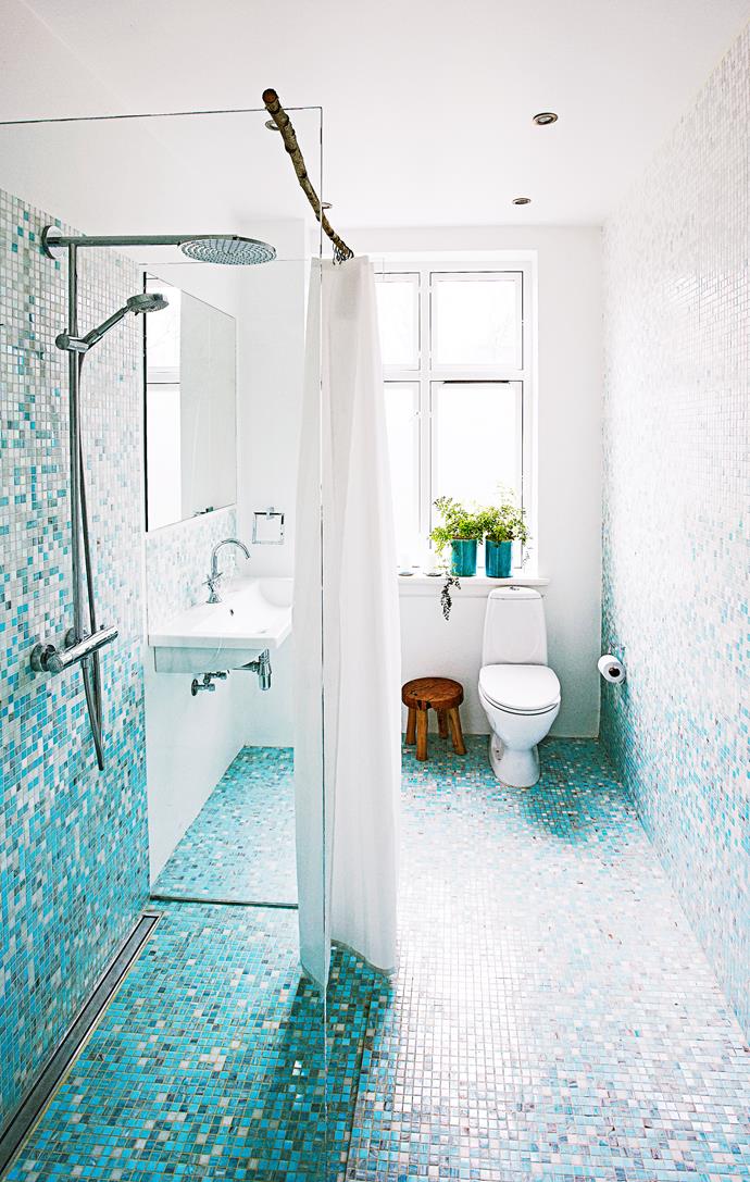 A tree branch is used as a shower curtain rod, an organic contrast in the crisp aqua mosaic-clad bathroom.