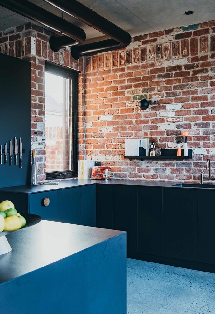 The kitchen space features black benchtops and sleek joinery that echo the black exposed pipes above and steel-framed windows. The simple design of the kitchen works seamlessly with the polished concrete flooring and red-brick walls, and the team have made clever use of space with a wall-mounted shelf for storage.
