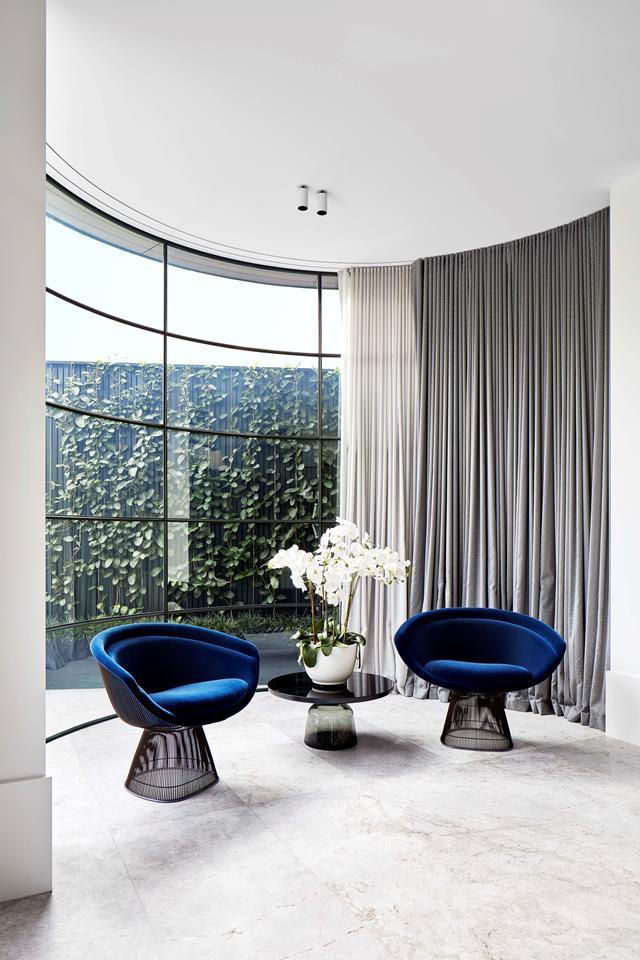 A black lacquer ClassiCon 'Bell' side table sits between a pair of Knoll 'Platner' chairs in blue velvet. The curved lines of the furniture echo the curved steel-framed window in this section of the living room.