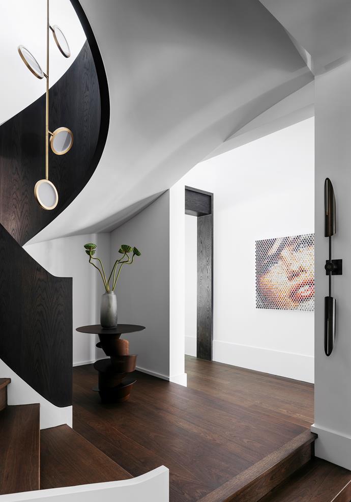 Custom 'Rachis' table by Dylan Farrell Design for Jean de Merry sits under the sweeping curve of the staircase which is clad in ebonised oak. Pendant light fitting is by Matter Made. Artwork is by Nemo Jantzen. The sconce is one of a pair by Allied Maker.