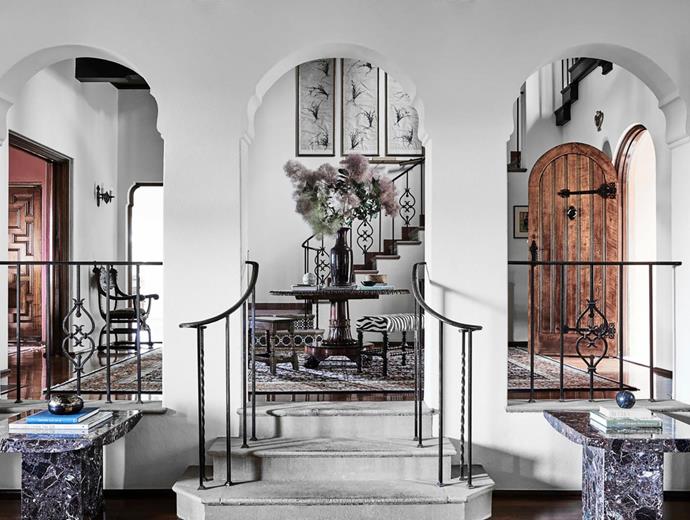 In the entry foyer, travertine marble side tables bookend the steps. Framed Chinese scrolls painted with Taoist poems hang above the stairs. An upholstered stool from The Vault and a late Regency mahogany table stand on a Persian Afshar-style rug. Floral arrangement by Mandalay Flowers.