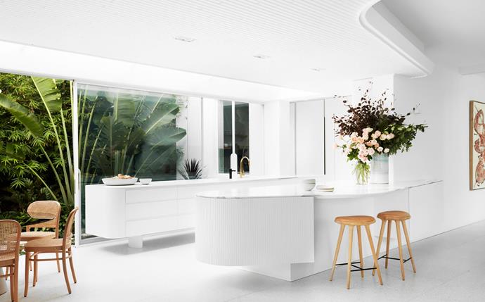 The bright white kitchen sits under a curved, timber-lined bulkhead, which is echoed in the shape of the bench topped with Statuario marble from Euro Marble. Astra Walker 'Icon' tapware from Candana. Mater oak and leather bar stools from Cult. Thonet 'N811' oak and rattan dining chairs.