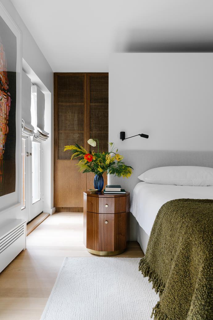 A custom bedhead in beige bouclé has been integrated into a  wall panel near ceiling height that creates an airiness and serves to separate the sleeping quarters from the walk-in robe and its back row of closets in this bedroom of this [warm and inviting update to a brownstone townhouse in New York City](https://www.homestolove.com.au/renovated-brownstone-townhouse-new-york-city-22805|target="_blank"). Tom Dixon bouclé bed throw in Khaki. 'Quillen' marquetry bedside table from Anthropologie. A Chuck Close artwork hangs near the windows, which are shaded with wool blinds. 
