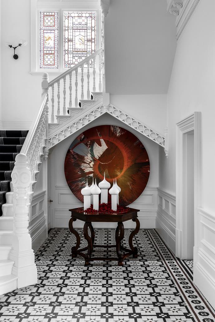 An artwork by Damien Hirst, Beautiful Romance in the Age of Uncertainty, hangs below the stairwell in the entry foyer. The A-frame above is a new steel support disguised by decorative detailing. Sculptures by Emily Floyd on a table by Marc French & Co. Original 1800s decorative floor tiles.