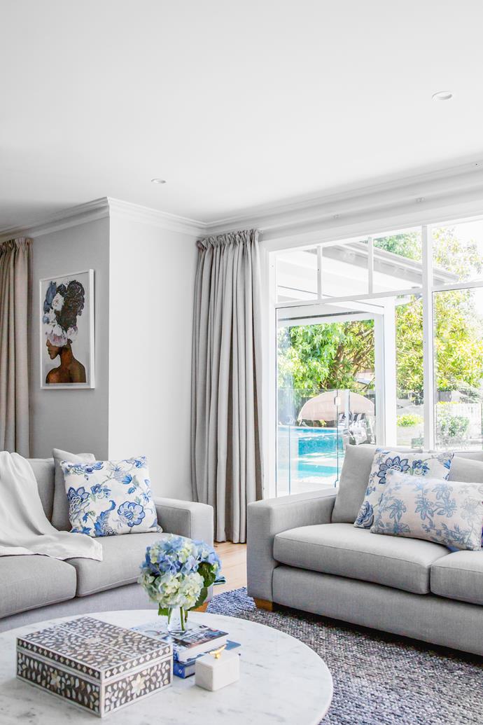 Wide windows welcoming natural light and a verdant outlook to the pool and garden are flanked by sumptuous custom-made floor-length curtains. "The curtains provide cosiness and add to the luxe feel," says interior-designer Belinda Vandenboom. Bespoke sofas by Furniture Innovations are scattered with floral pillows that reflect the blue peonies in Brent Rosenberg's Everything I Am print. The walls throughout are painted in Dulux White Exchange Quarter.