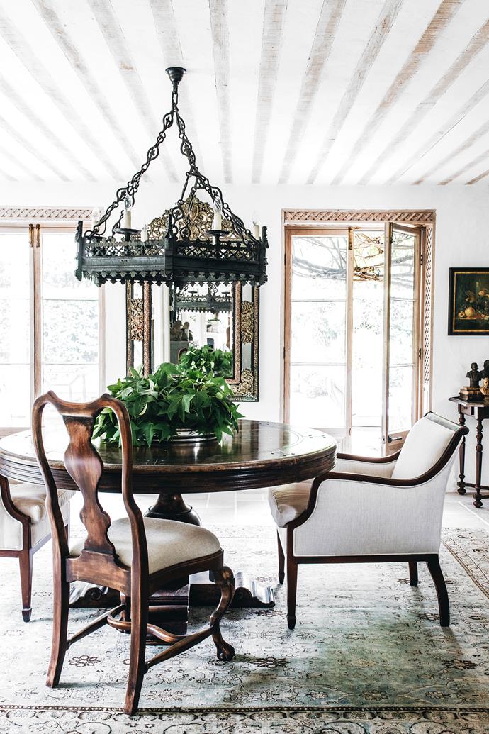 A statement light fitting over the dining table serves as a focal point, not only of the room but also of the view from adjoining rooms. A c1750 French forged iron hexagonal pendant light with Gothic detailing hangs above a dark-stained timber table with scroll bracket feet. Upholstered club chair in shaded walnut finish and Swedish Louis XVI dining chair in polished timber. C1860 French repoussé brass-mounted cushion mirror.