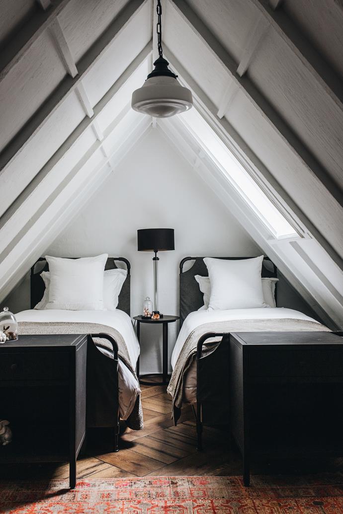 In an attic bedroom with a vaulted ceiling and dormer windows, a Parisian architectural milk-glass brasserie pendant light hangs above 'French Académie' iron panel beds with footboards and 'French Directoire' glass column lamp, all from RH.