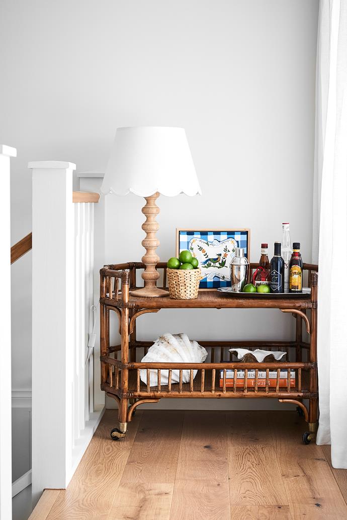 In a corner, the cane bar cart was designed by Maine House Interiors, who also created the scalloped shade, which sits on a lamp base from Provincial Home Living. The artwork is by [Emma Sheehan](https://www.emmasheehanartist.com/home|target="_blank"|rel="nofollow").