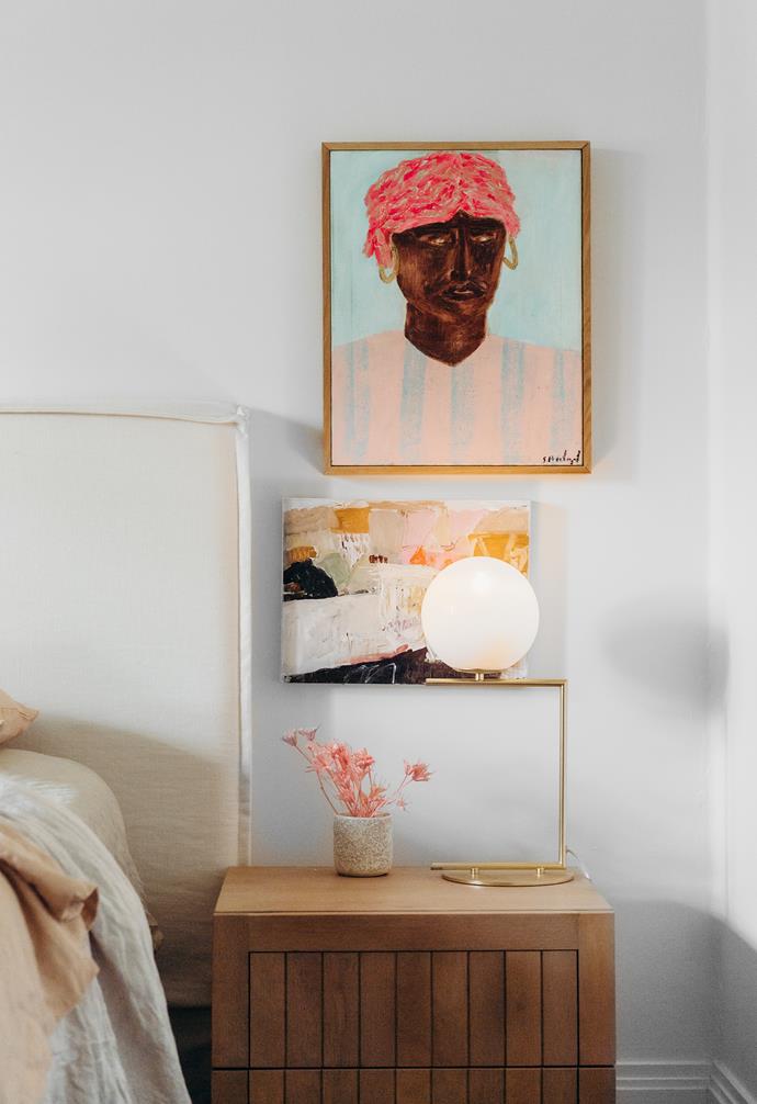 A gold lamp from Adairs sits atop a timber bedside table from GlobeWest in the master bedroom. On the wall, a portrait by Stanislas Piechaczek and an abstract landscape by Giorgia Bel bring colour and character. The lined bedhead is from Create Estate.