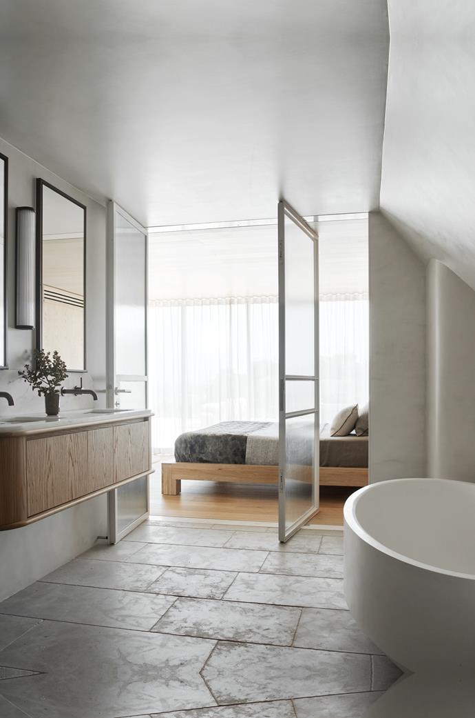 Large pivot steel-and-glass doors intimately connect the bedroom of this [elegant penthouse in Tamarama](https://www.homestolove.com.au/penthouse-renovation-tamarama-sydney-24911|target="_blank") to its lavish ensuite bathroom. Tumbled Garonne limestone floor tiles and custom microcemented walls and ceiling give the space a luxe-spa vibe.